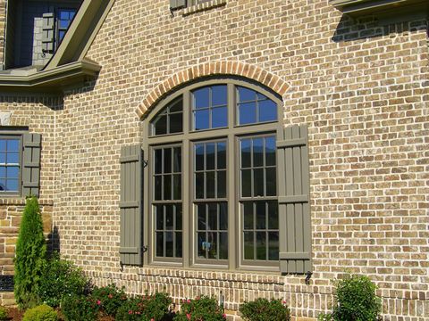 Olive open board and batten shutters flanking an arched window on a brick home.