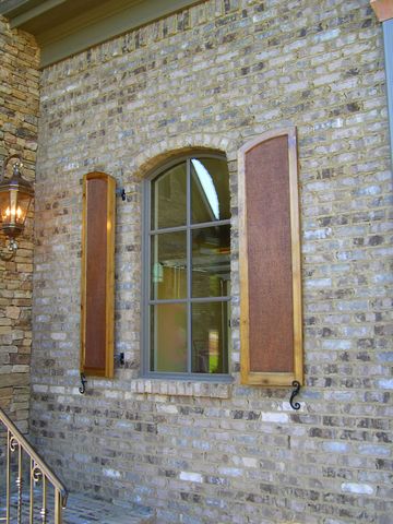 Stained arch-top flat panel shutters with rough cedar panels beside an arched window, brick and stone.