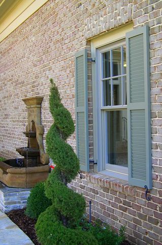 Sage Charleston louvered shutters on a brick home, with a spiral topiary at the corner.
