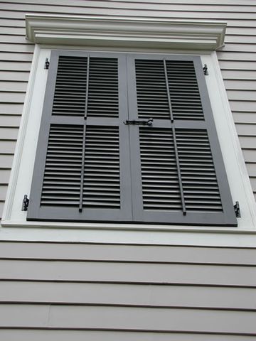 Black louvered shutters closed over a window on lap siding, showing the hinges and slide bolt.