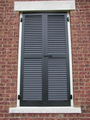 Black louvered shutters closed over a window in red brick, with a slide bolt.