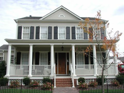 Tan two-story home with black louvered shutters and a wrap-around porch, fall color.