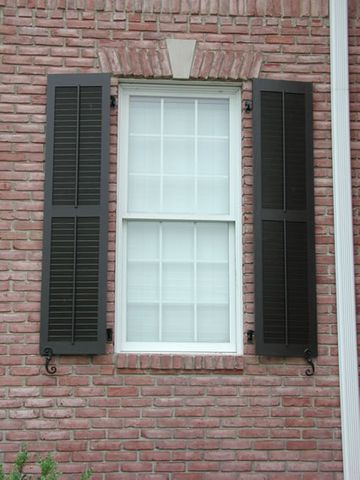 Black louvered shutters on brick, hung on hinges with S-scroll shutter dogs.