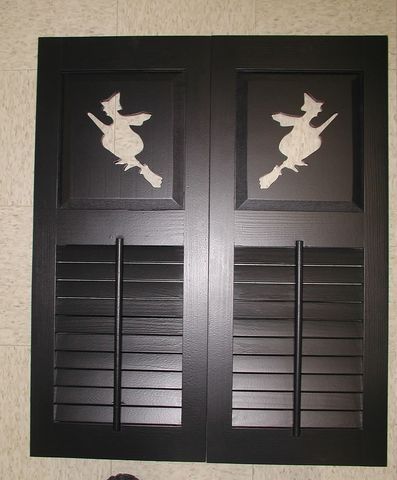 Black louver-over-flat-panel shutters with witch-on-broom cutouts in the top panels.