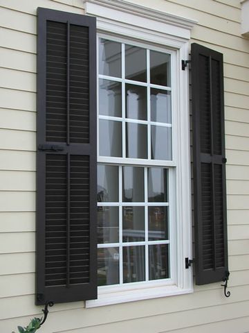 Dark louvered shutters on a yellow clapboard home, hung on hinges with shutter dogs.