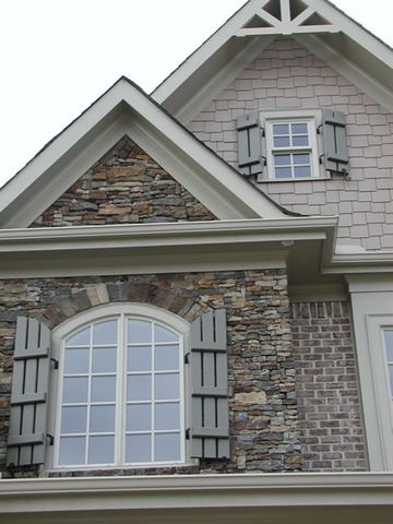 Sage open board and batten shutters on the gable windows of a brick and stone home.