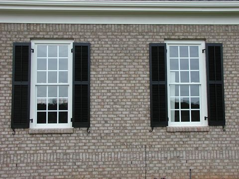 Black louvered shutters on tan brick, hung on hinges with S-scroll shutter dogs.