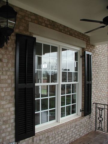 Black louvered shutters on a tan brick porch window, with a shutter dog.