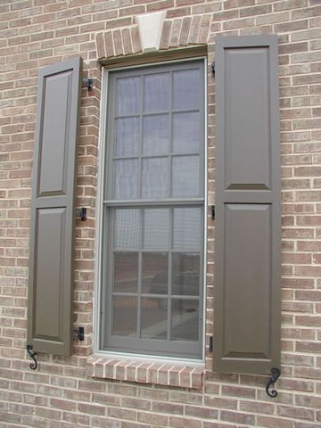 Dark brown flat panel shutters on tan brick, hung on hinges with S-scroll shutter dogs.