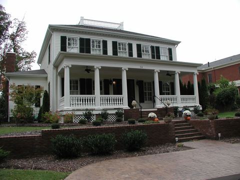 White two-story with black louvered shutters and a deep columned porch, fall decorations.