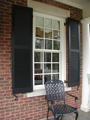 Black louvered shutters on red brick beside a porch, hung on hinges with shutter dogs.