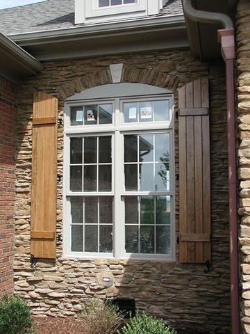Natural stain-grade board and batten shutters flanking an arched window in stacked stone.