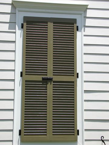 Olive louvered shutters closed over a window on white siding, with hinges and a slide bolt.