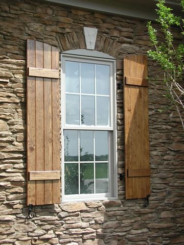 Natural stain-grade closed board and batten shutters on a stacked-stone wall, with shutter dogs.