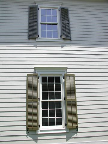 Olive louvered shutters on grey lap siding, hung on hinges with shutter dogs.