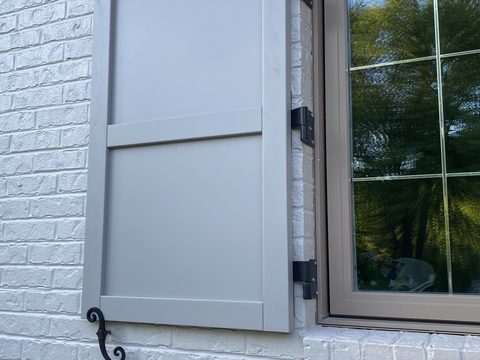 Light gray composite flat panel shutter on white-painted brick, with black hinges and an S-scroll shutter dog.
