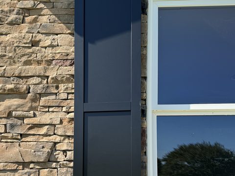 Black composite flat panel shutter on stacked stone, with a white-framed window.