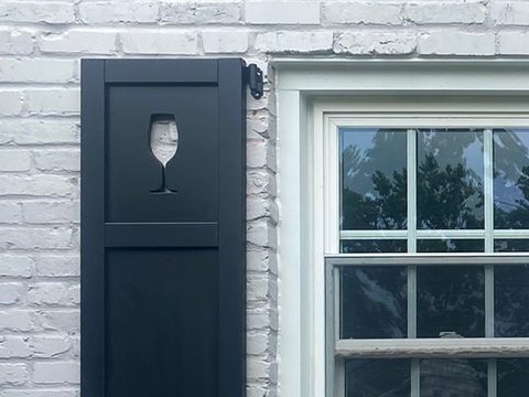Black composite flat panel shutter with a custom wine glass cutout in the top panel, on white-painted brick with black hinges and an S-scroll shutter dog.