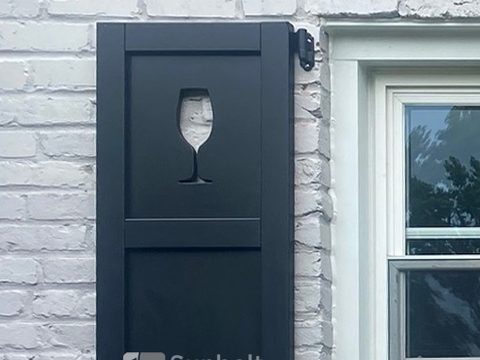 Close view of a black composite flat panel shutter with a custom wine glass cutout and black hinge.