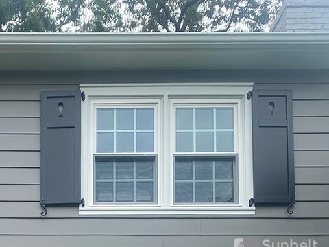 Charcoal composite flat panel shutters with wine glass cutouts flanking a double window on gray lap siding, with S-scroll shutter dogs.