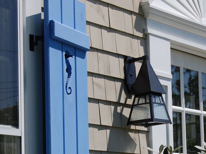 Blue custom board and batten shutter with a seahorse cutout beside a lantern on a shingled coastal cottage