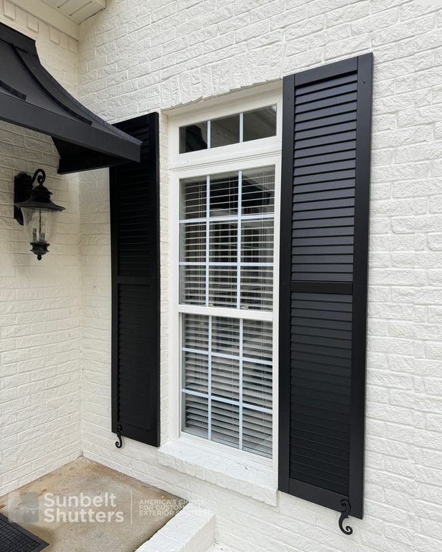 Black custom louvered shutters beside a window on a white brick home with a lantern