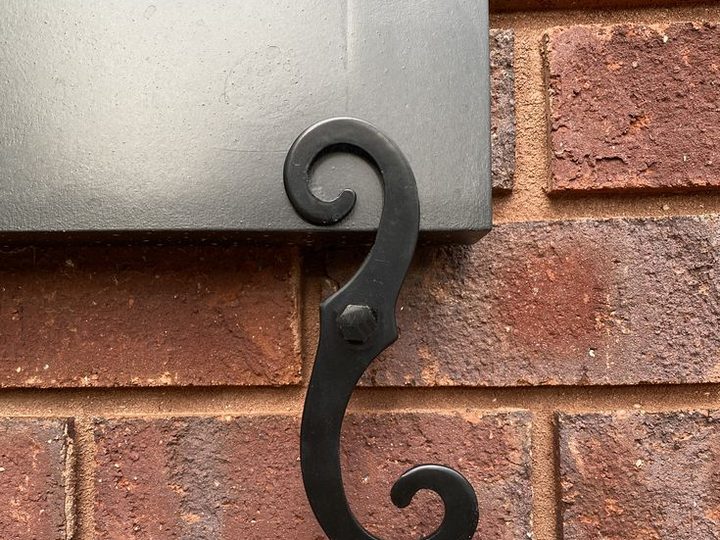 Black S-scroll shutter dog tieback mounted on a red brick wall beside a gray shutter